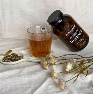 Brown jar with herbal tea with a label "Defence" by Welb Organics, leaning against a cup of tea on a small plate with dried herbs next to a bunch of flowers.