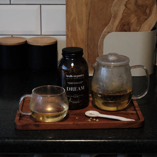 Brown jar of herbal tea labelled "Welb Organics Dream" on a wooden tray next to a glass and a tea pot.