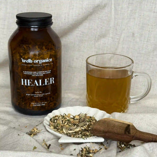 Brown jar of herbal tea with a label "Welb Organics Healer" next to a cup of tea and a mix of blended herbs and wooden spoon.