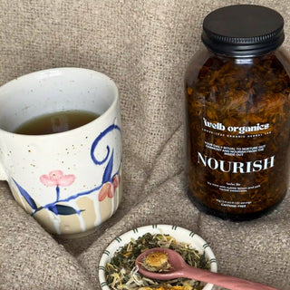 Tea mug with floral design, jar of tea labeled 'Nourish', and spoon with dried herbs on a textured surface.