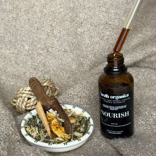 Brown glass bottle of herbal tincture  labeled 'Nourish' with a dropper, next to a small dish with herbs on a textured surface.