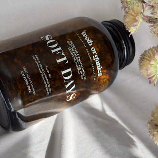 Bottle of Welb Organics 'Soft Days' product on a light surface with flowers in the background