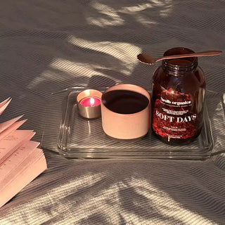 A styled flat-lay scene on a grey striped fabric surface with dappled shadows. A clear glass tray holds a pink ceramic cup, a small lit pink candle in a silver holder, and a dark amber bottle labeled 'welb organics SOFT DAYS' with a spoon resting on top.