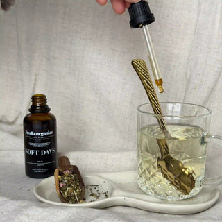 Bottle of herbal tincture labeled 'soft days' with a dropper over a glass of liquid on a neutral background