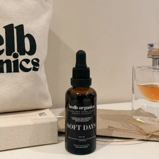A dark amber glass dropper bottle labeled 'welb organics SOFT DAYS' stands on a tabletop, styled with a cream canvas tote bag, a book, and a perfume bottle in the background.